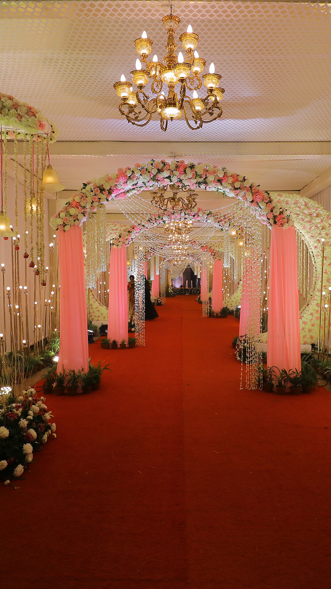 Grand wedding entryway with pink floral arches, red carpet aisle, crystal chandeliers, and fairy light décor